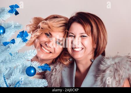 Elegant girlfriends in evening dresses and a fur collar stand nearby. Sisters hug. Young beautiful women smiling in anticipation of Christmas, gatheri Stock Photo