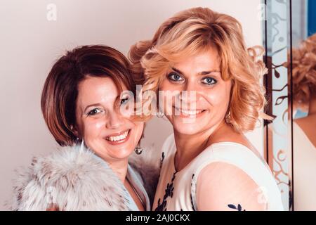 Elegant girlfriends in evening dresses and a fur collar stand nearby mirror. Sisters hug. Young beautiful women smiling in anticipation of Christmas, Stock Photo