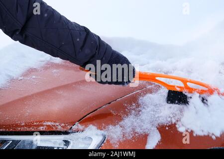 Snowy. Brush in mans hand. Man clears orange car from snow. Car covered with snow. Winter problems of car drivers. Stock Photo
