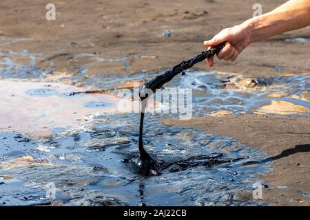 A man's hand stirring liquid asphalt with a wooden stick at Pitch Lake, the largest natural deposit of bitumen in the world. La Brea, Trinidad island, Stock Photo