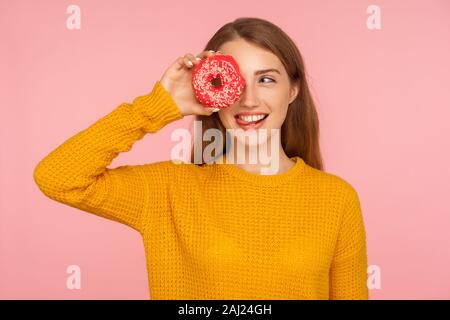 Portrait of playful red hair girl in sweater looking through hole in doughnut and sticking out tongue, peeking into pink donut, having fun with sweet Stock Photo