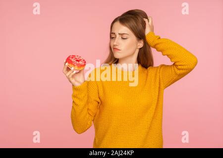 Portrait of confused ginger girl in sweater scratching head and hesitating to eat sweet doughnut, wants sugary donut for snack and doubting about junk Stock Photo