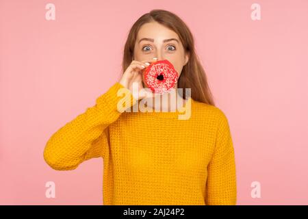 Portrait of positive red hair girl in sweater covering mouth with doughnut and looking amazed at camera, putting pink donut on lips, having fun with s Stock Photo