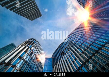 Modern high-rise buildings bottom view. Sun shine on blue sky. Top part of skyscrapers in city center. Sunbeams mirroring on facade. Metropolis detail. Stock Photo