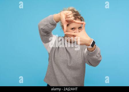 Photographer view. Curious woman in grey sweatshirt pretending to take picture, focusing on camera through photo frame hand gesture, looking with pryi Stock Photo