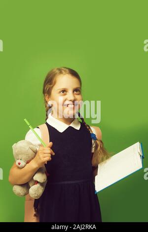 Girl with braids and happy face. Pupil holds blue book, marker and teddy bear. Kid in school uniform isolated on green background. Study and back to school concept. Stock Photo