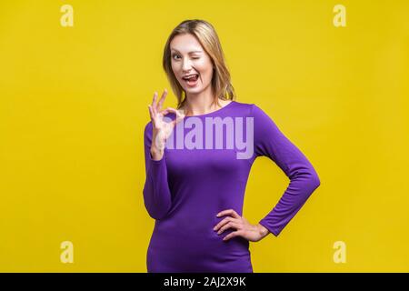 Everything is fine. Portrait of playful positive woman in purple tight dress standing with one hand on hips, winking at camera and showing ok gesture. Stock Photo