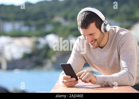 Happy adult man with headphones checking smart phone content in a balcony on the beach Stock Photo
