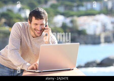 Happy adult man using laptop and talking on smart phone in a balcony on the beach Stock Photo