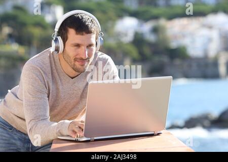 Serious man wearing wireless headphones uses a laptop in a balcony on the beach Stock Photo