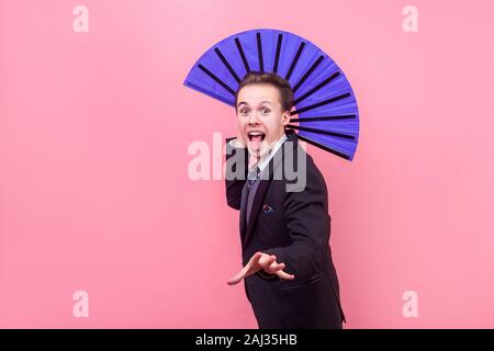 Portrait of joyous carefree young man, magician in elegant suit sticking out tongue and holding big blue fan, illusionist showing performance, having Stock Photo