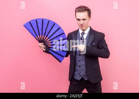 Portrait of young positive magician, man in elegant suit standing pointing at big blue fan, showing trick performance and looking with intriguing expr Stock Photo