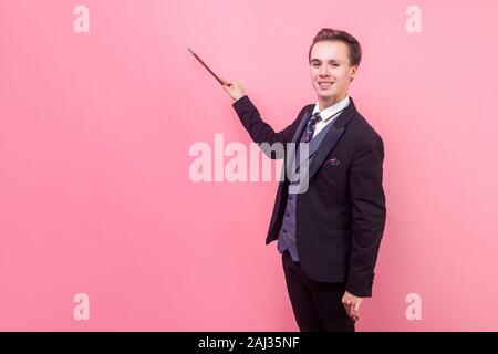 Portrait of happy positive magician in tuxedo standing with raised magic wand showing trick, smiling at camera, illusionist performance. indoor studio Stock Photo