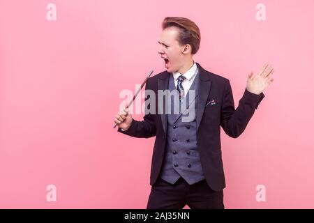 Portrait of positive illusionist with magic wand or male artist in tuxedo making announcement, pretending to sing with magic stick as a microphone. st Stock Photo