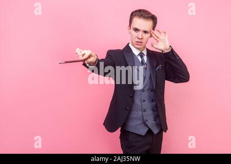 Portrait of concentrated focused magician in tuxedo touching temple with fingers and raising magic wand, using mental power to perform trick, illusion Stock Photo