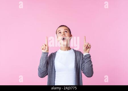 Look above! Portrait of amazed brunette teen girl with bun hairstyle in casual clothes standing with open mouth in surprise, pointing up at copy space Stock Photo