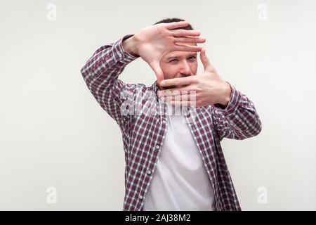 Portrait of young man in casual plaid shirt standing looking at camera through frame made of fingers, capturing photo, focusing on distant with curiou Stock Photo