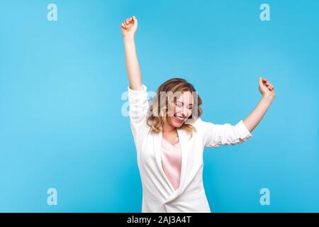 I'm winner! Portrait of ecstatic overjoyed lady with wavy hair in white jacket dancing with raised arms, smiling excitedly, celebrating victory, succe Stock Photo