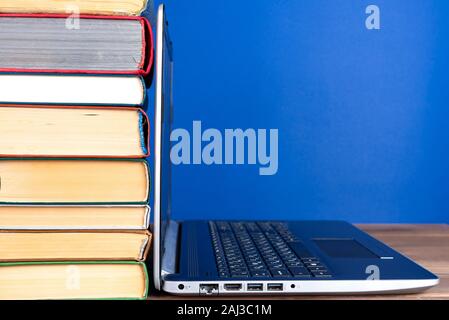 laptop next to a stack of colorful books on a wooden table on a blue background Stock Photo