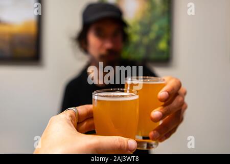 two beer glasses cheering close up selective focus point of view, a caucasian man wearing black hat in the bar background, beer tasting room Stock Photo