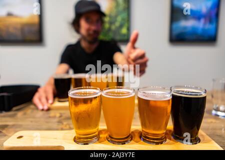 selective focus Flight of Craft of four different Beers glasses on Wooden Tray, happy caucasian man giving thumb sign finger gesture in the background Stock Photo