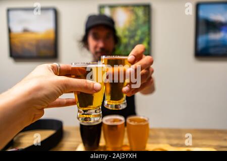 two beer glasses cheering close up selective focus point of view, man in the bar background, flight of Craft of different Beers glasses on Wooden Tray Stock Photo