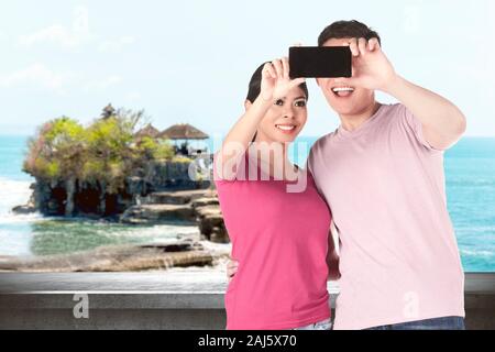 Asian couples making a selfie using a camera phone with Tanah Lot Temple background Stock Photo
