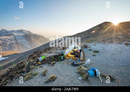 Man pokes head out of tent at sunset while camping on mountain ridge. Stock Photo