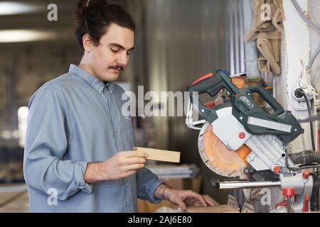 Young carpenter looking at wooden detail and examining it while standing standing in front of electric saw in workshop Stock Photo