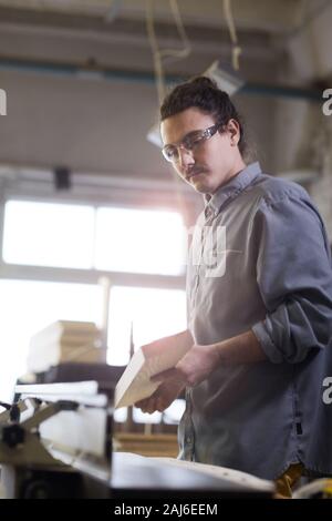 Young man in protective eyeglasses standing and holding wooden detail and working on lathe in the plant Stock Photo