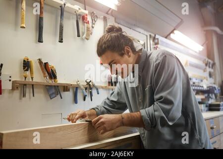 Young carpenter standing at his workplace and grinding the wooden plank with special equipment in workshop Stock Photo