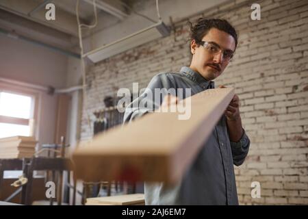 Young carpenter in protective eyeglasses examining the quality of wooden detail while working in the plant Stock Photo