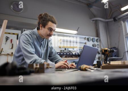 Young repairman working online he typing on laptop computer at the table in workshop Stock Photo