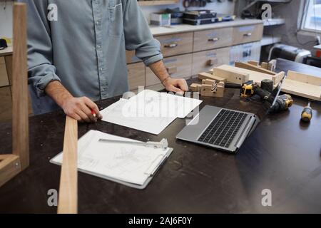 Close-up of architect standing at the table with laptop and work tools and examining blueprint in workshop Stock Photo