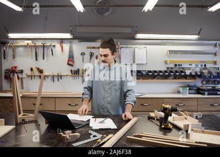 Young carpenter using laptop at the table while working with wooden plank in workshop Stock Photo