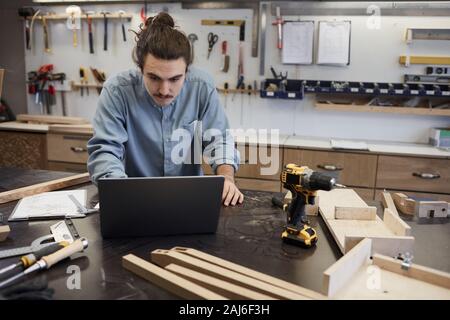 Young carpenter using laptop while standing at the table with wooden details he looking at online pattern and working with wood in workshop Stock Photo