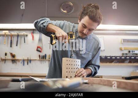 Young man making holes with drill in wooden details while working in workshop Stock Photo