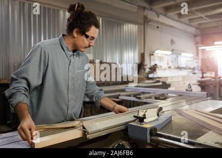 Young carpenter in protective eyeglasses standing at lathe and working with wooden details in workshop Stock Photo