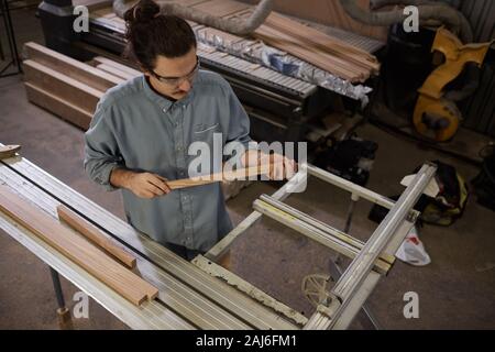 Young carpenter examining the wooden plank in his hands while working on lathe in workshop Stock Photo