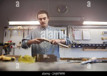 Young man standing near his workplace and fastening two wooden planks he making things from wood in workshop Stock Photo