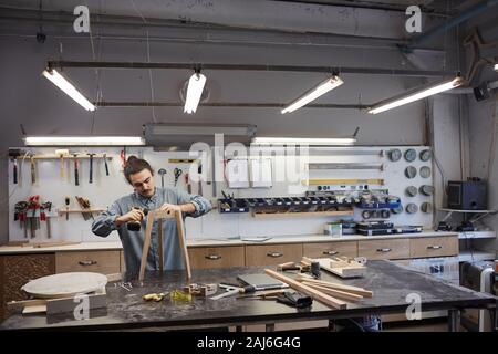 Carpenter standing near the workplace with wooden chair on it and using drill in his work in workshop Stock Photo