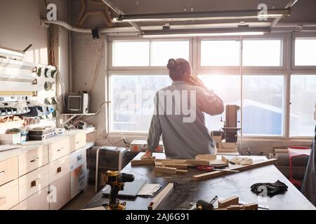 Rear view of carpenter sitting on the table and talking on mobile phone he taking order while working in workshop Stock Photo