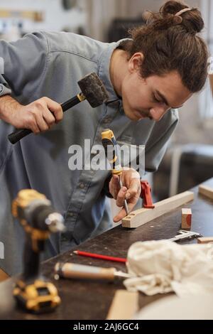 Young serious carpenter making holes on wooden plank using screwdriver and hammer in workshop Stock Photo
