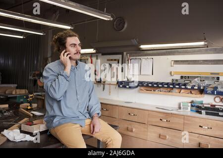 Young carpenter in casual clothing sitting on the table and has a conversation on mobile phone during his break in workshop Stock Photo