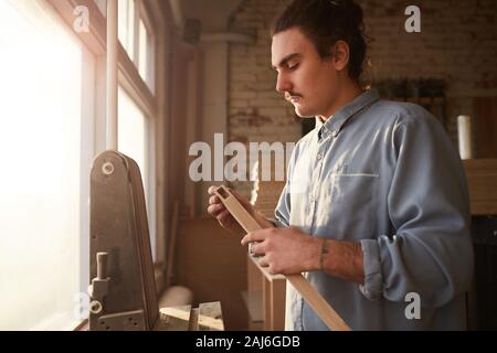 Serious manual worker standing in front of the lathe and examining the wooden plank Stock Photo