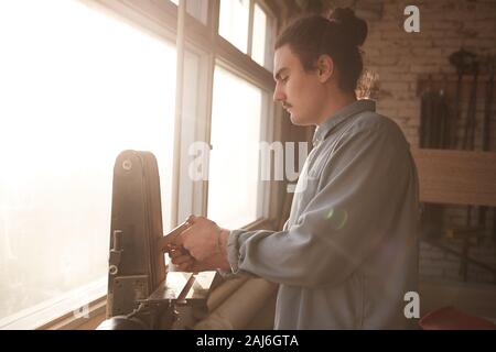 Side view of young carpenter standing in front of the window with sunlight and using lathe machine in his work in factory Stock Photo