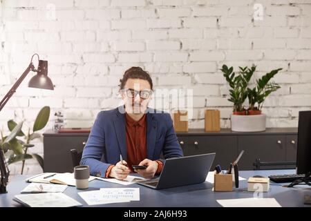 Young designer in eyeglasses looking at camera while working at the table with mobile phone and laptop at office Stock Photo