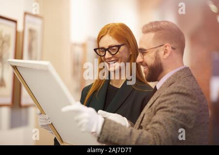Young woman in eyeglasses holding picture and looking at it together with young man they standing and smiling in gallery Stock Photo
