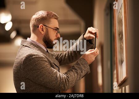 Side view of young bearded man in eyeglasses taking photo of paintings on his mobile phone in the gallery Stock Photo