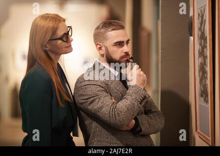 Young couple in formal wear standing together in front of the painting on the wall and enjoying the art in the gallery Stock Photo